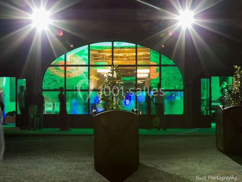 Façade vitrée éclairée de nuit avec des silhouettes visibles à l'intérieur et des projecteurs lumineux à l'extérieur.