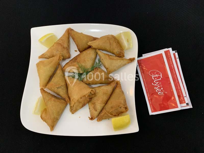 Assortiment de samoussas dorés sur une assiette blanche, accompagnés de quartiers de citron et de sachets de sauce.