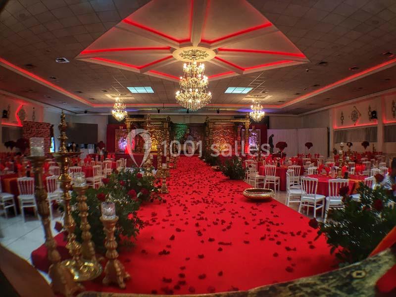 Salle de réception décorée avec un tapis rouge, chandeliers dorés, pétales de rose et tables dressées.