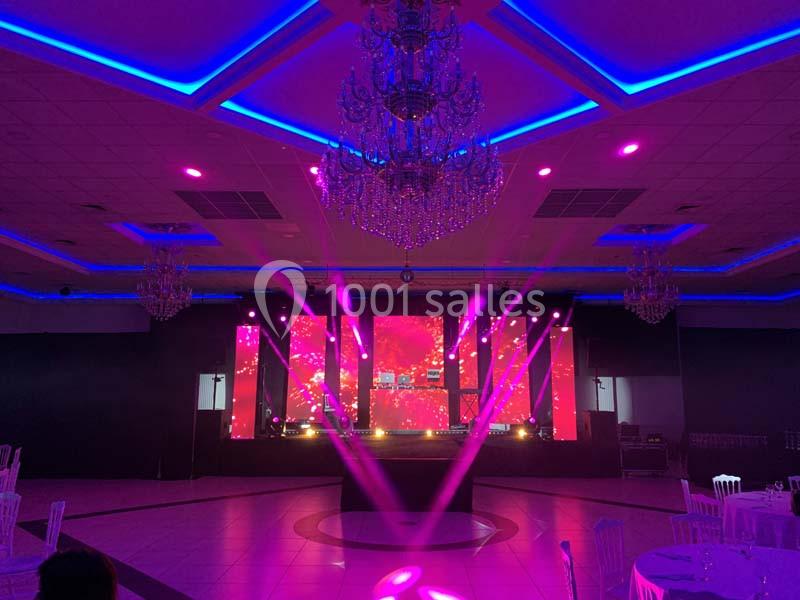 Salle de réception illuminée par des projecteurs roses, avec une scène équipée d'écrans LED et des lustres suspendus.