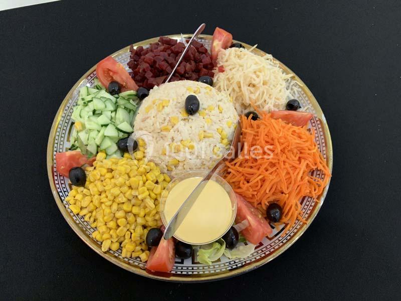 Assiette composée de riz au maïs, crudités variées, olives noires et sauce servie dans un petit récipient.