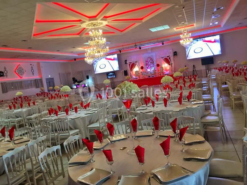 Salle de réception décorée avec des tables rondes, nappes blanches, serviettes rouges et éclairage lumineux.