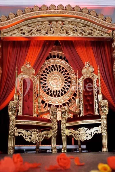 Deux trônes dorés ornés de motifs complexes, placés devant un décor rouge et or avec un grand motif circulaire.