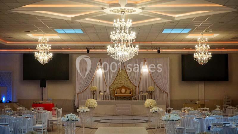 Salle de réception élégante avec lustres, tables décorées de fleurs blanches et une estrade ornée de rideaux et d'un canapé.