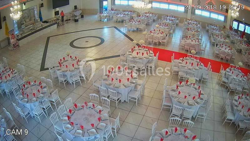 Salle de réception spacieuse avec des tables rondes dressées, nappes blanches et serviettes rouges, éclairage lumineux.