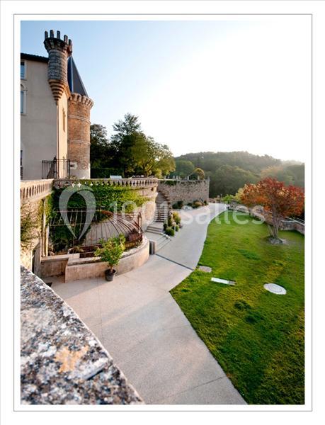 Chemin pavé longeant un château avec tour, jardin verdoyant et vue dégagée sur la campagne environnante.