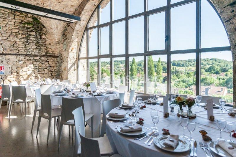 Salle de réception lumineuse avec grandes baies vitrées, tables rondes dressées et vue sur un paysage verdoyant.