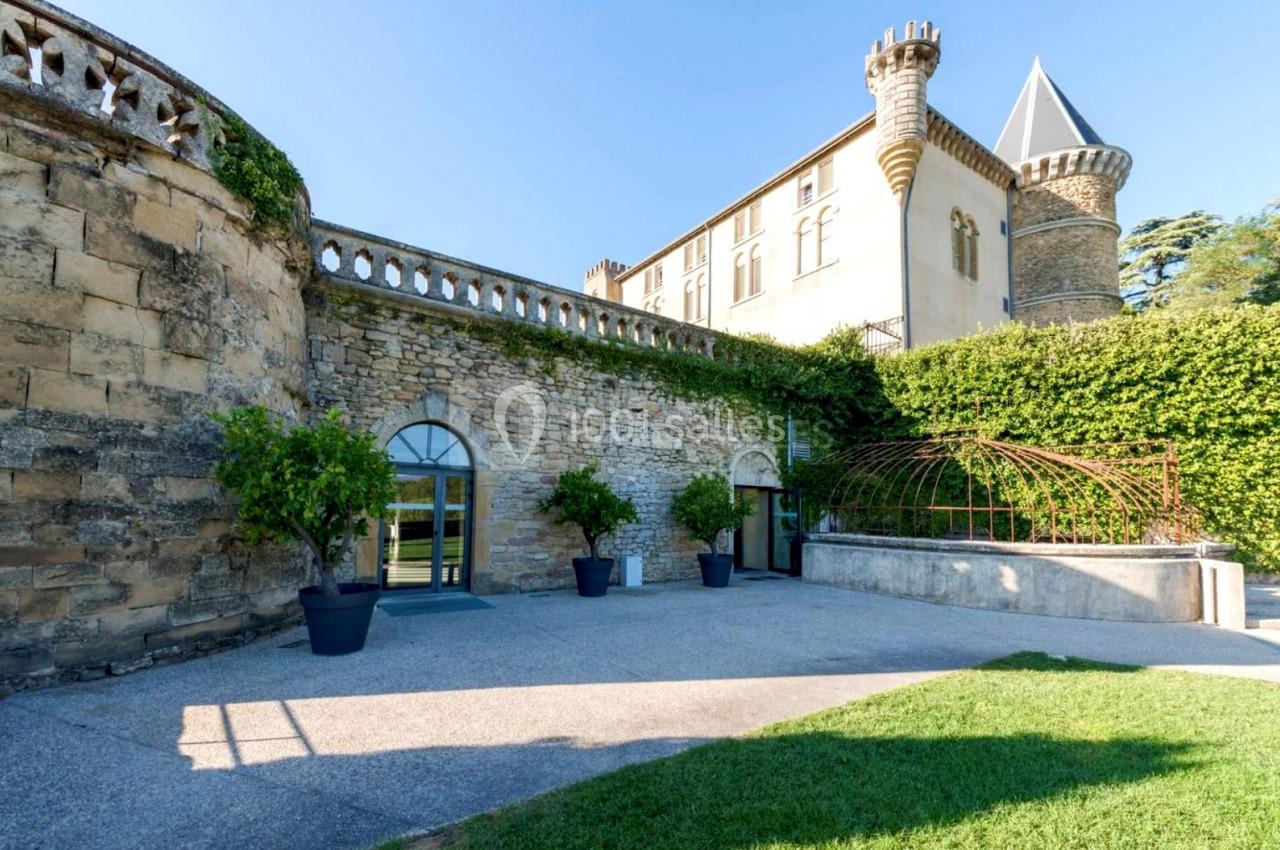 Façade en pierre d'un château avec tours, grandes fenêtres et terrasse bordée de verdure.