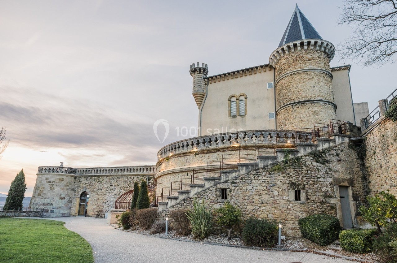Château en pierre avec tours rondes et escalier extérieur, entouré d'un jardin et d'un chemin pavé.