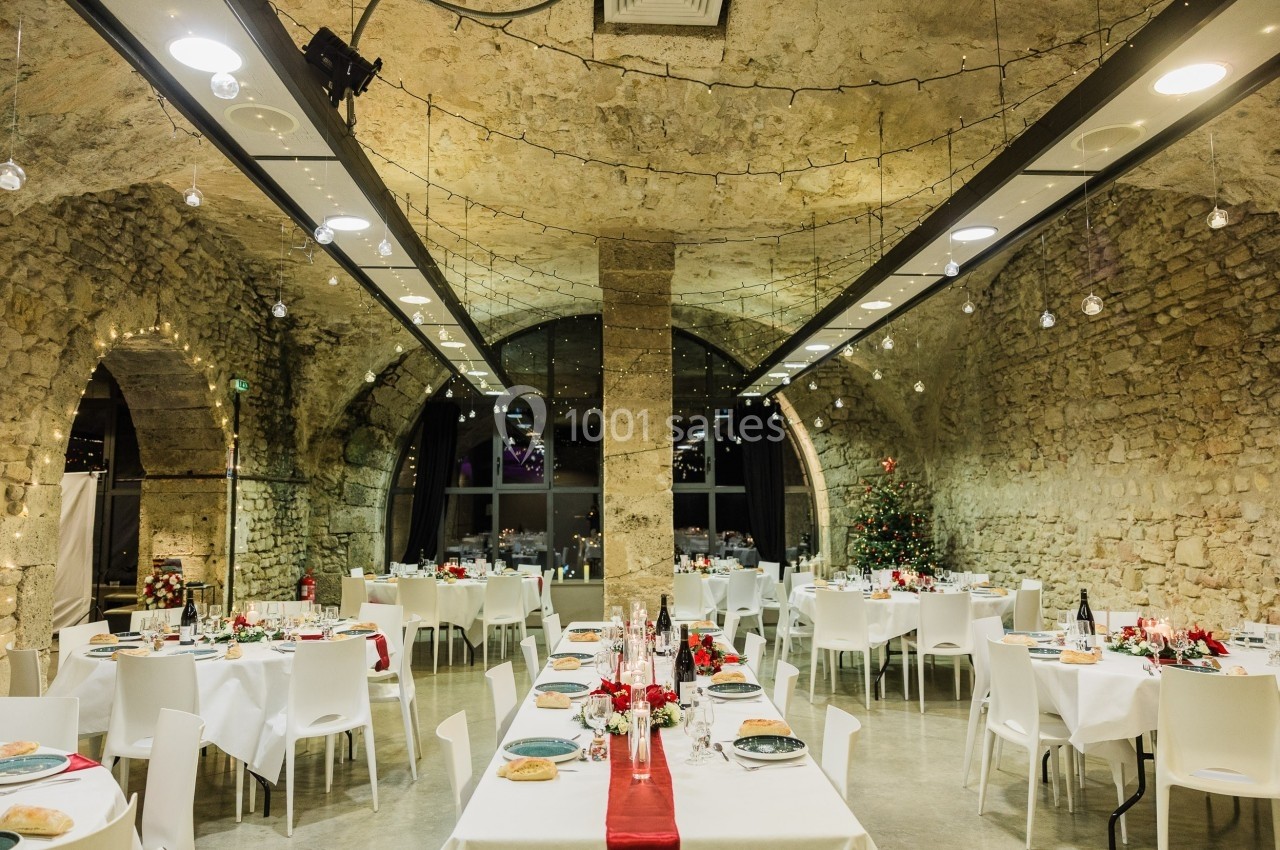 Salle en pierre voûtée décorée pour un repas festif, avec tables dressées, nappes blanches et guirlandes lumineuses.