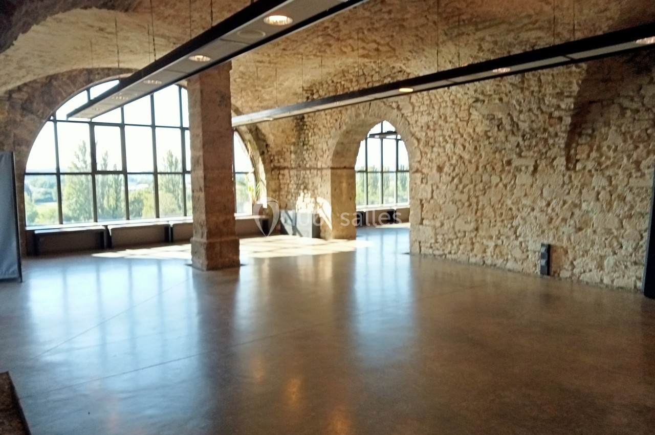 Salle spacieuse avec murs en pierre, grandes arches et baies vitrées laissant entrer la lumière naturelle.