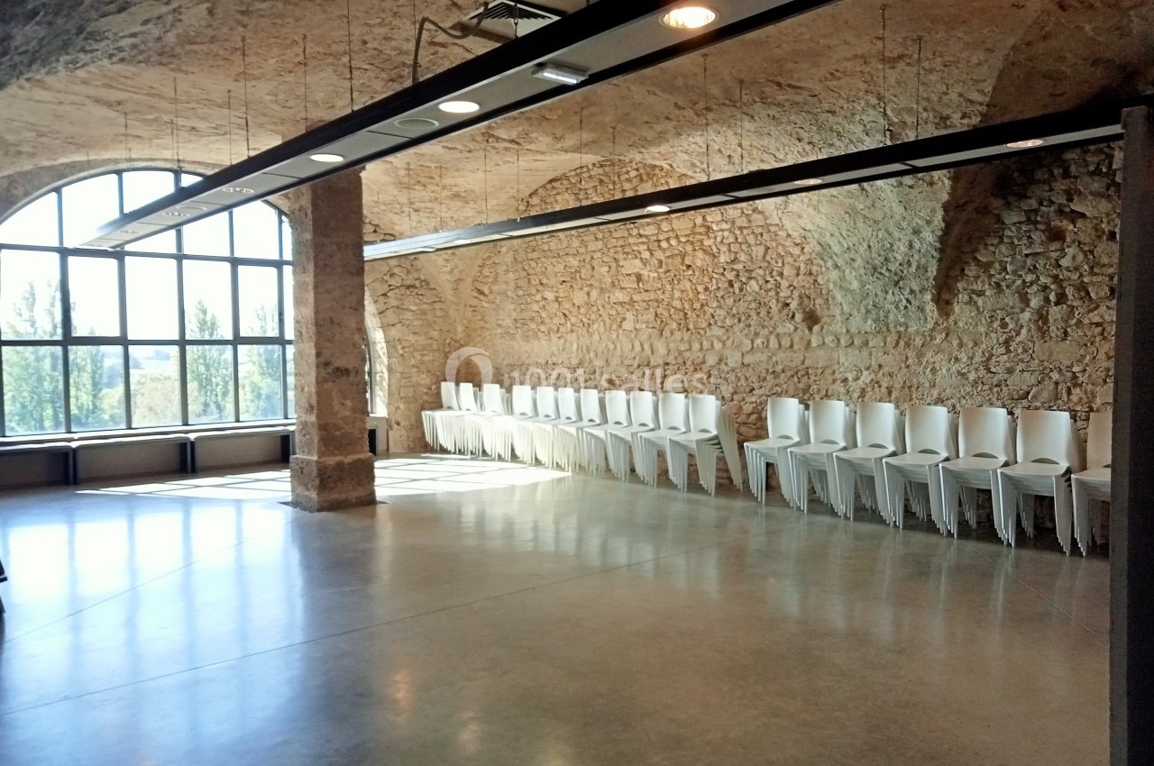 Salle lumineuse avec murs en pierre, grandes fenêtres cintrées et rangée de chaises blanches alignées contre un mur.
