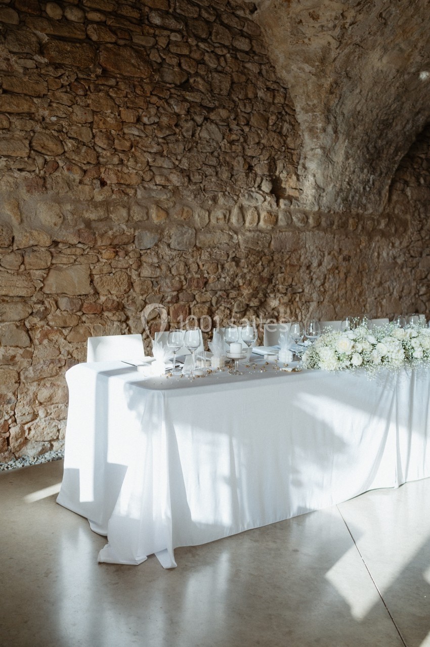 Table de réception élégamment dressée avec nappes blanches, vaisselle raffinée et décor floral, dans une salle en pierre.