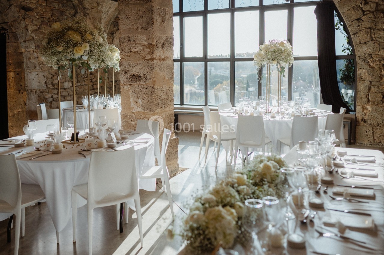 Salle de réception lumineuse avec tables décorées de fleurs blanches et jaunes, entourées de chaises blanches.