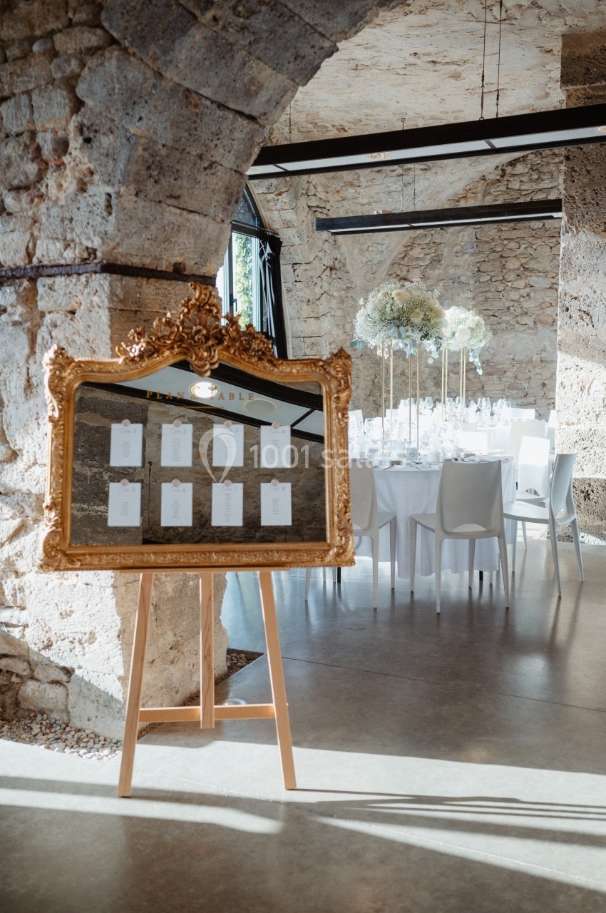 Plan de table encadré en or sur un chevalet, devant une salle de réception décorée avec des tables et des fleurs blanches.