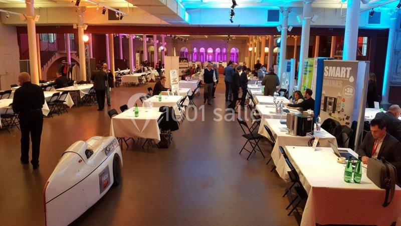 Salle d'exposition avec stands, visiteurs et une voiture futuriste blanche exposée au premier plan.