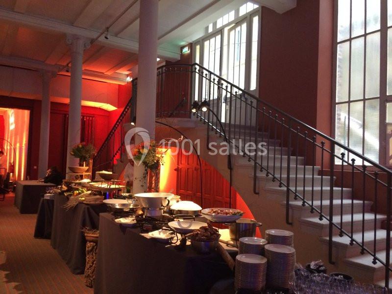 Buffet dressé dans une salle lumineuse avec escalier, colonnes blanches et décorations florales.