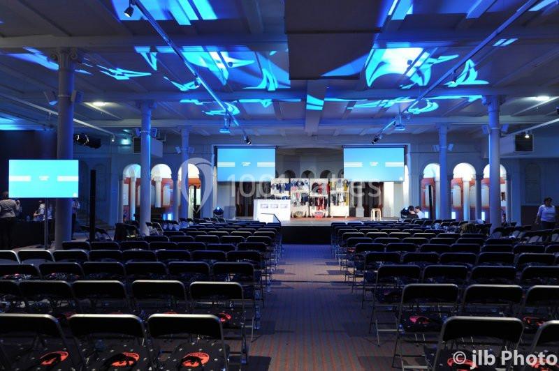 Salle de conférence vide avec des rangées de chaises, une scène éclairée en bleu et des écrans de présentation.