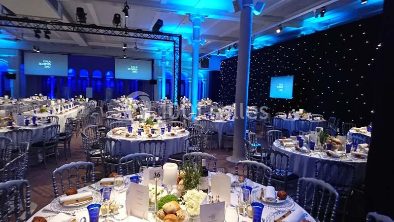 Salle de réception élégamment décorée avec des tables rondes dressées, éclairage bleu et écrans en arrière-plan.