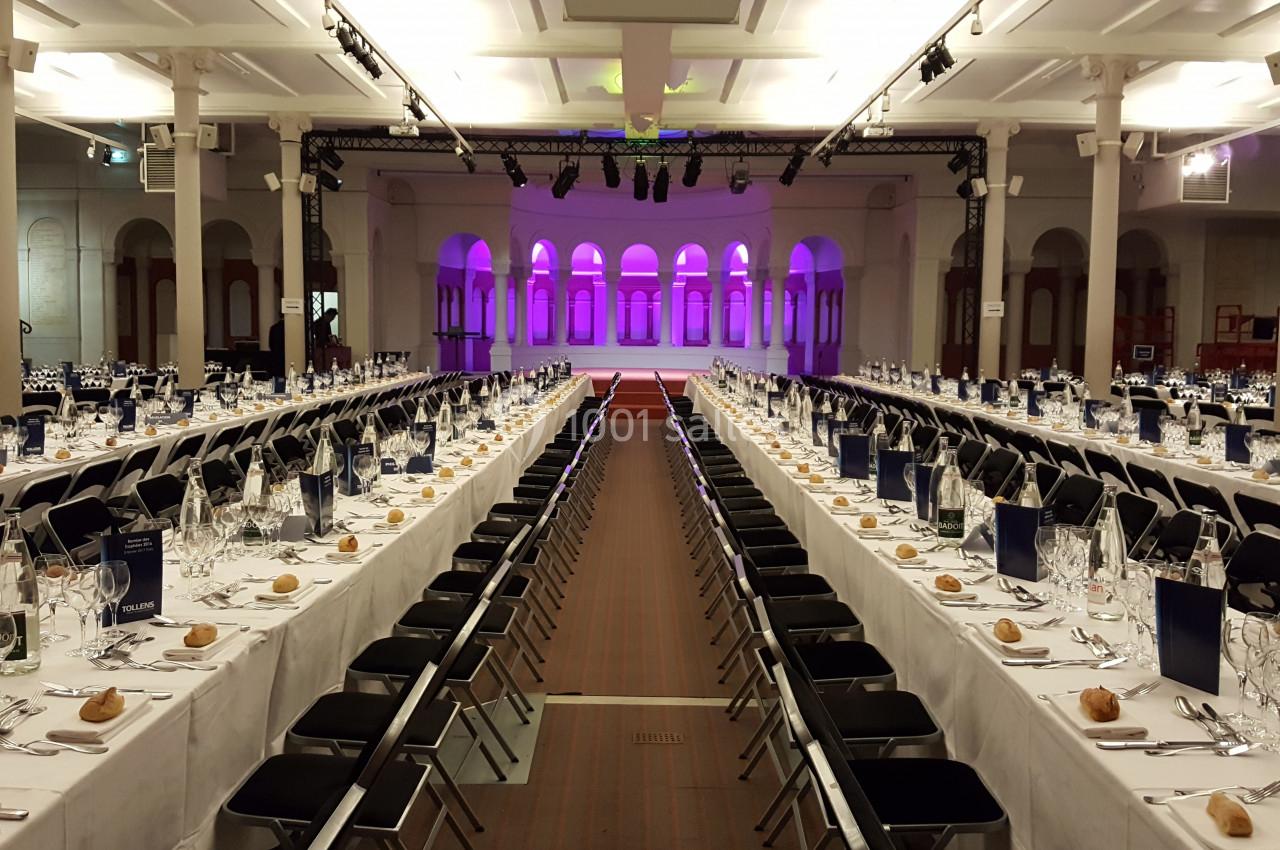 Salle de réception aménagée avec de longues tables dressées pour un banquet, éclairage violet en arrière-plan.