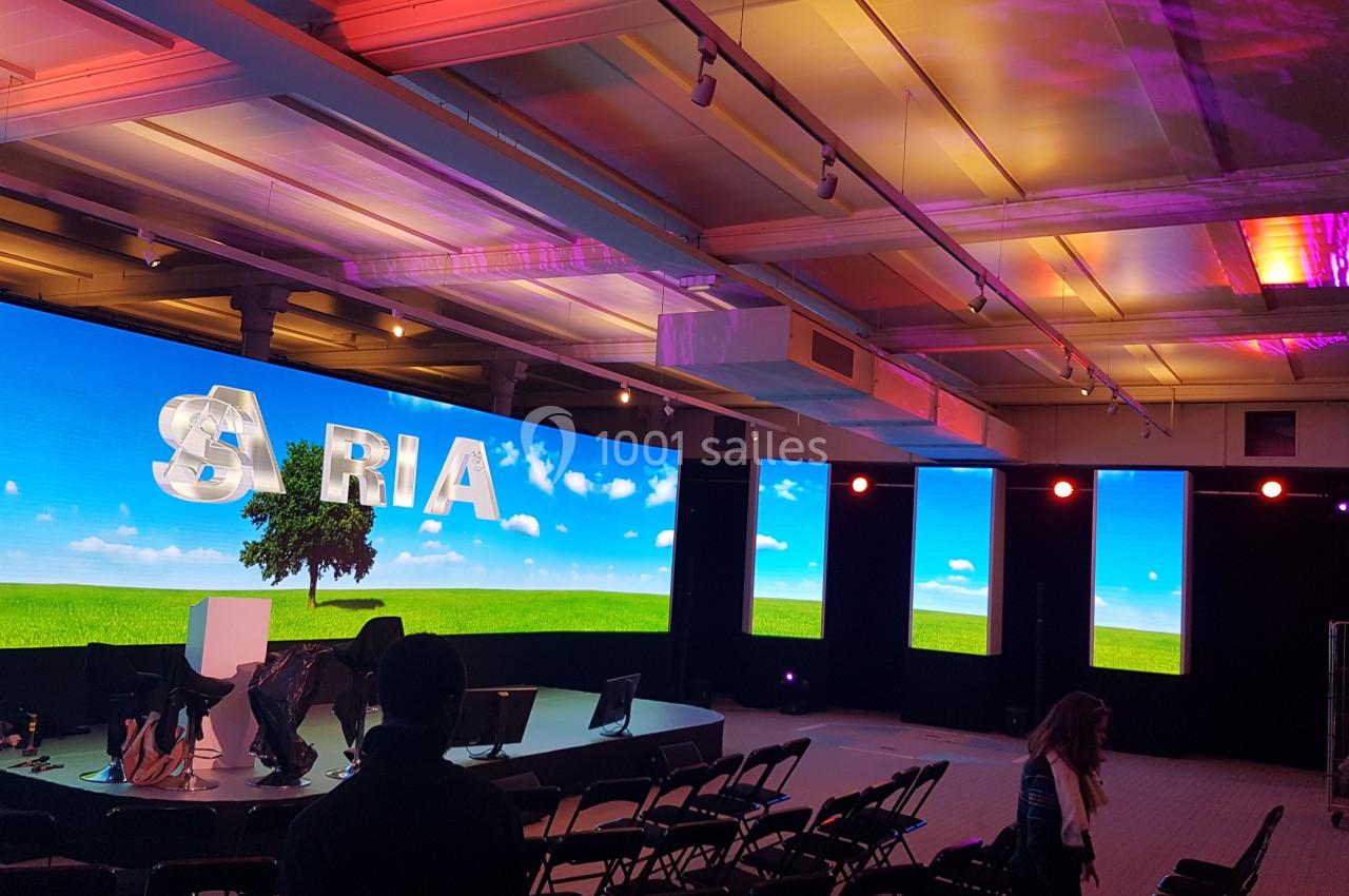 Salle de conférence avec écrans géants affichant un paysage champêtre et le mot ’ARIA’, éclairage coloré.