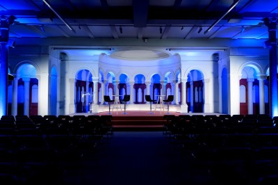 Salle de conférence vide avec rangées de sièges rouges, écran de projection et éclairage fluorescent.