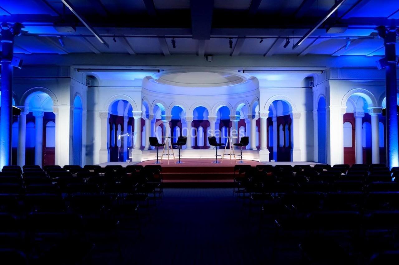 Salle de conférence avec scène éclairée en bleu, trois chaises hautes et rangées de sièges vides au premier plan.