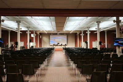 Salle de conférence vide avec rangées de sièges rouges, écran de projection et éclairage fluorescent.