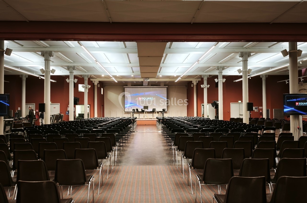 Salle de conférence vide avec des rangées de chaises noires, un écran principal et deux écrans latéraux allumés.