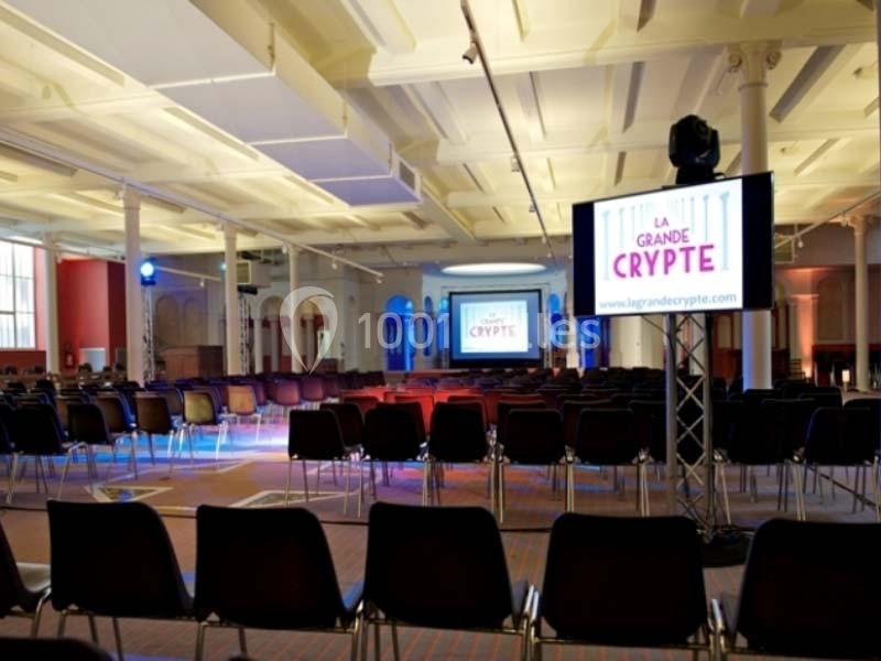 Salle de conférence lumineuse avec des rangées de chaises vides et un écran affichant ’La Grande Crypte’.