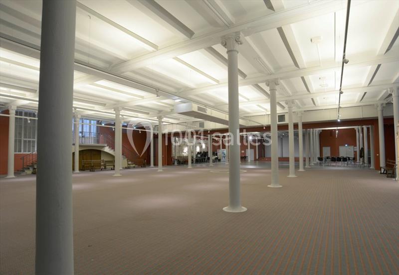 Grande salle vide avec plafond haut, colonnes blanches, moquette beige et murs rouges, éclairée par des lumières…