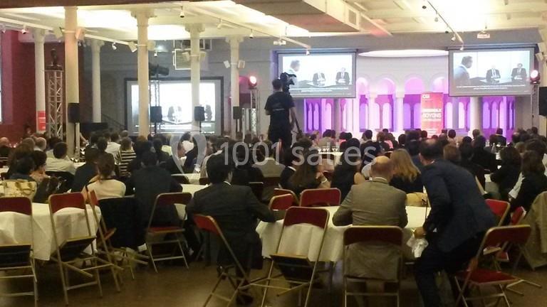 Salle de conférence avec un public assis, un caméraman et des écrans diffusant une présentation sur scène.