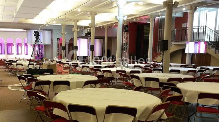 Salle de réception avec des tables rondes dressées, des chaises rouges et un éclairage tamisé.