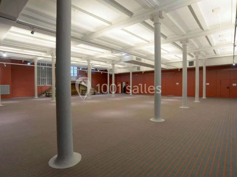 Salle spacieuse avec colonnes blanches, murs rouges et sol beige, éclairée par des lumières au plafond.