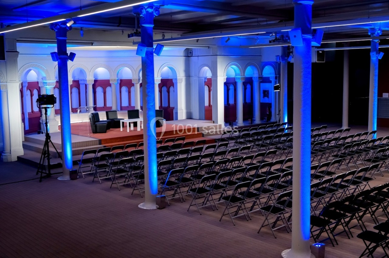 Salle de conférence vide avec des rangées de chaises noires, une scène éclairée et des colonnes blanches.