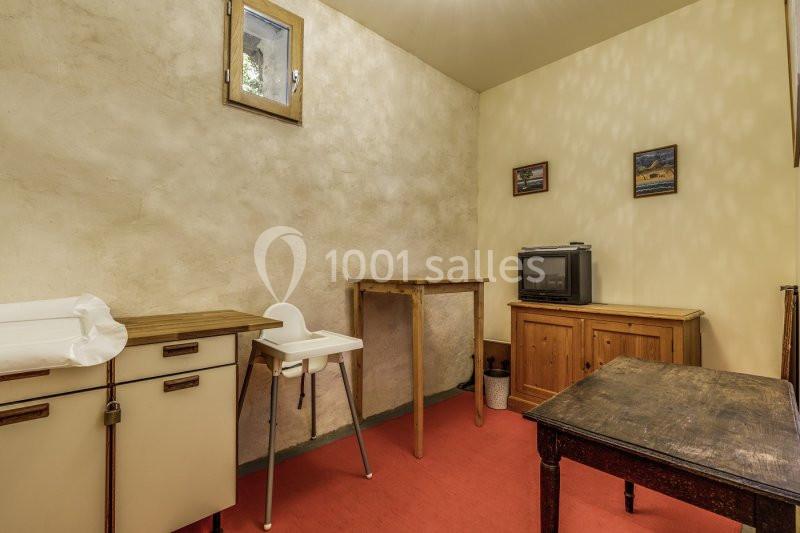 Salle simple avec murs beige, sol rouge, table en bois, chaise haute, meuble bas et petite télévision.