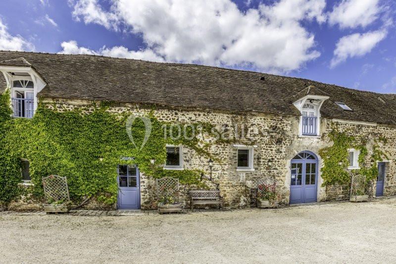 Façade en pierre d'une maison ancienne couverte de lierre, avec portes et fenêtres bleues sous un ciel partiellement nuageux.