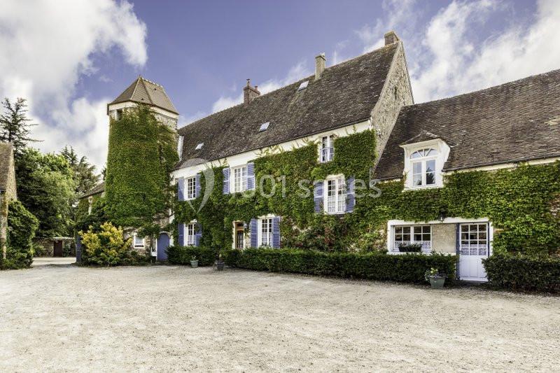 Façade d'une grande maison en pierre recouverte de lierre, avec une cour en gravier et un ciel partiellement nuageux.