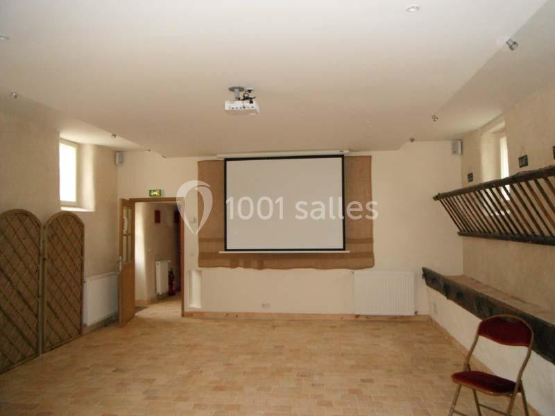 Salle vide avec un écran de projection, des murs clairs, un sol en carrelage et quelques chaises disposées sur les côtés.