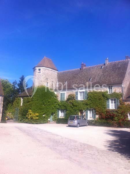 Grande maison en pierre avec tour, volets verts et façade partiellement couverte de lierre, sous un ciel bleu.
