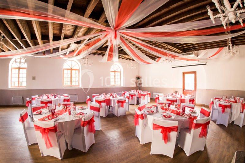 Salle de réception décorée de nappes blanches et rubans rouges, avec des tables rondes et des chaises assorties.