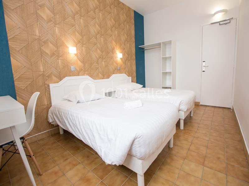 Deux lits simples avec draps blancs dans une chambre lumineuse, décorée avec un mur texturé et un sol en carrelage.