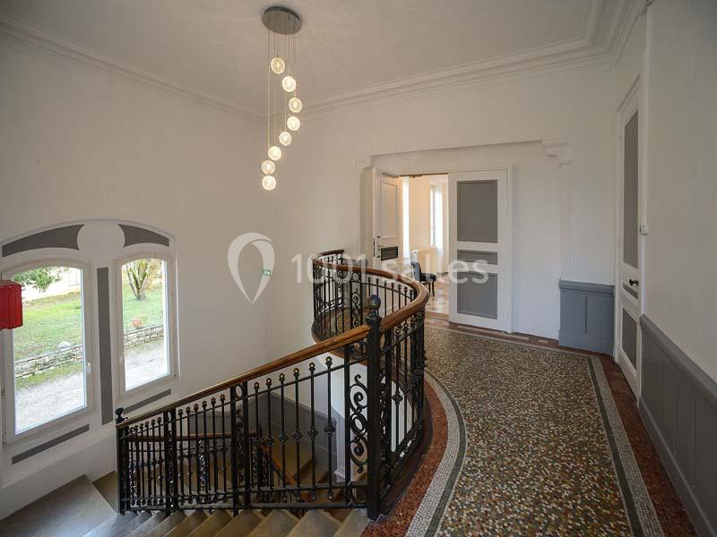 Escalier intérieur en colimaçon avec rampe en fer forgé, sol en mosaïque et lumière naturelle provenant des fenêtres.