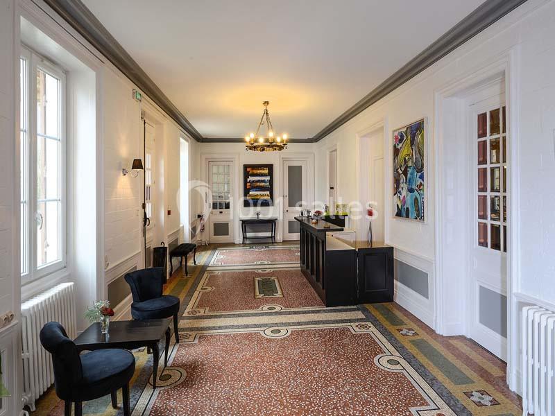 Couloir lumineux avec sol en mosaïque, murs blancs ornés de tableaux, mobilier noir et fenêtres laissant entrer la lumière.