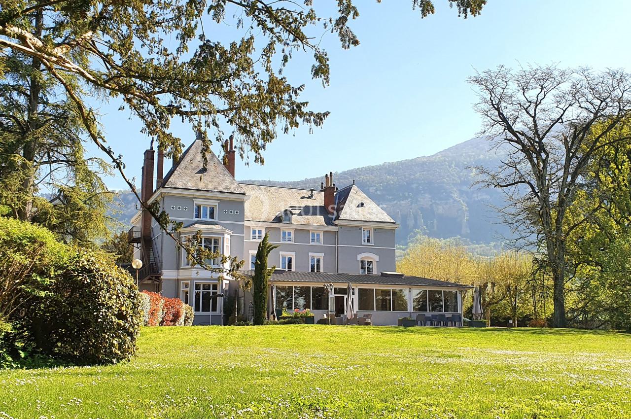 Manoir entouré de verdure avec des arbres et des montagnes en arrière-plan sous un ciel dégagé.