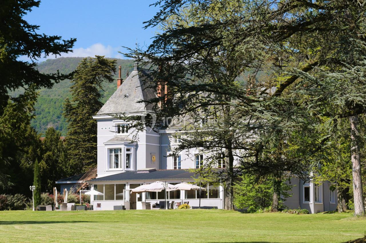 Manoir blanc entouré d'arbres et d'une pelouse, avec terrasse et parasols, au pied de collines verdoyantes.
