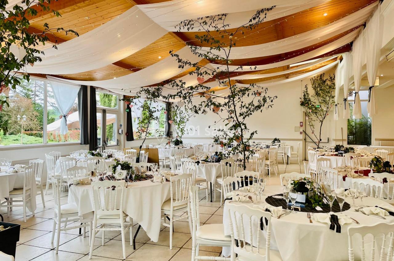 Salle de réception décorée avec des tables rondes, nappes blanches, chaises blanches et branches végétales suspendues.