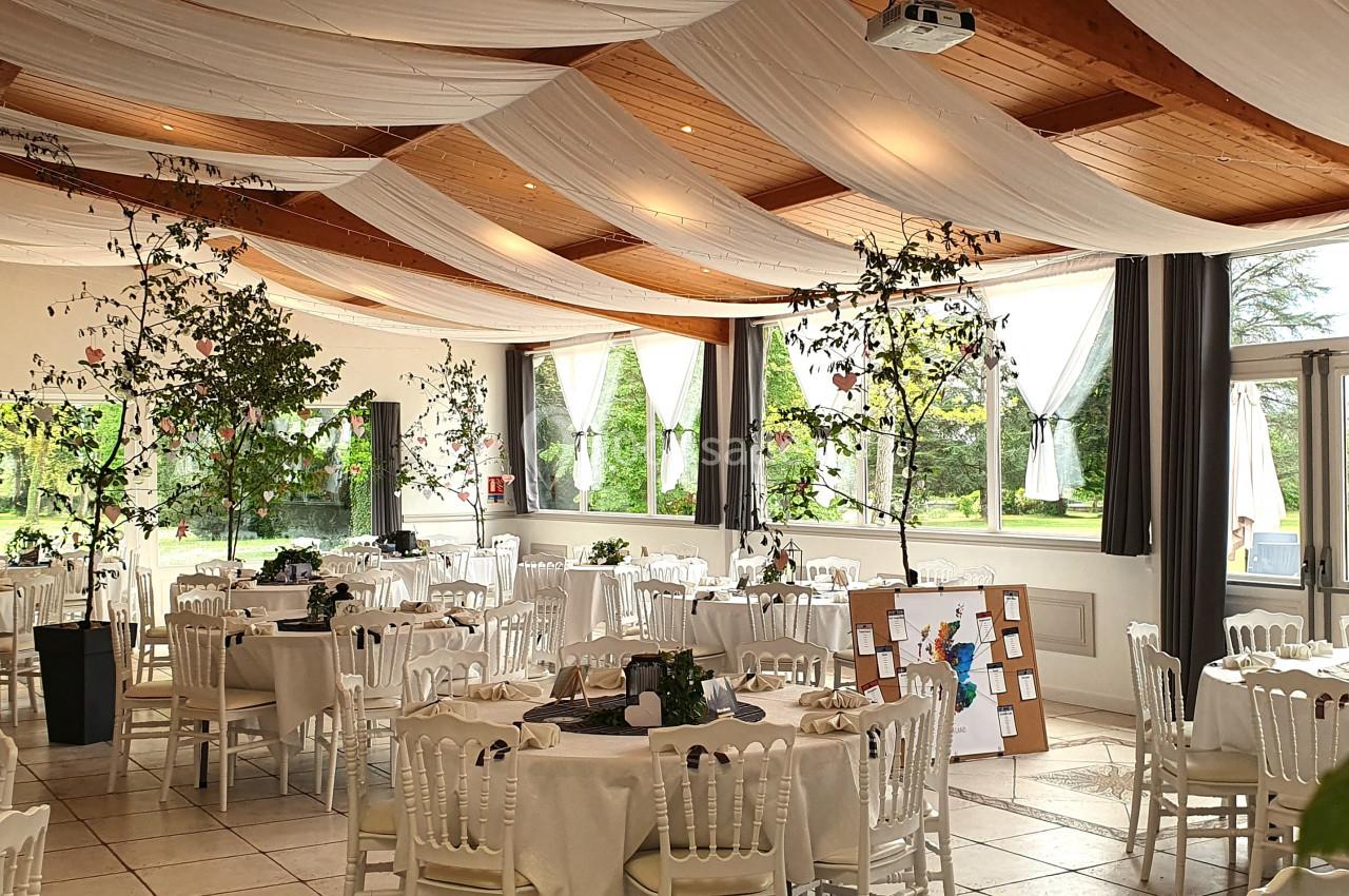 Salle de réception lumineuse avec tables rondes dressées, chaises blanches et décorations végétales suspendues.
