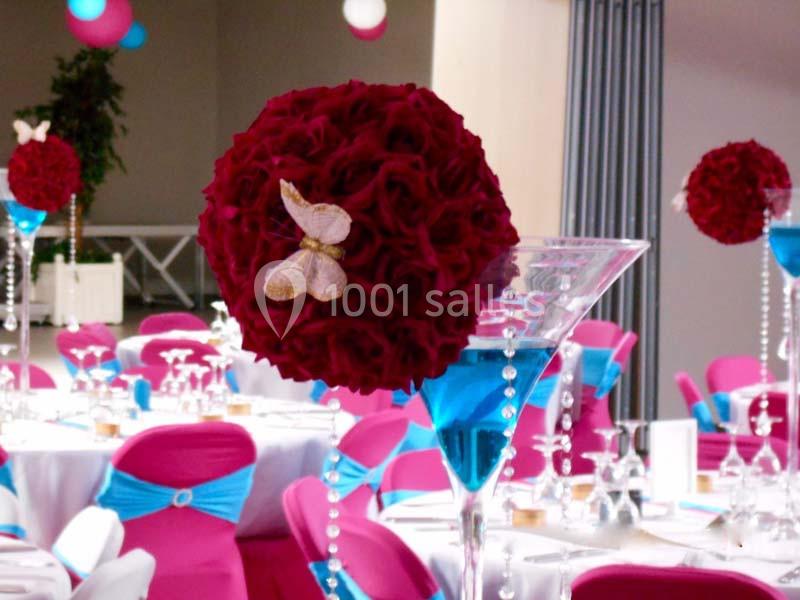 Centre de table avec boule de roses rouges, papillon décoratif et vase rempli d'eau bleue, entouré de chaises roses.