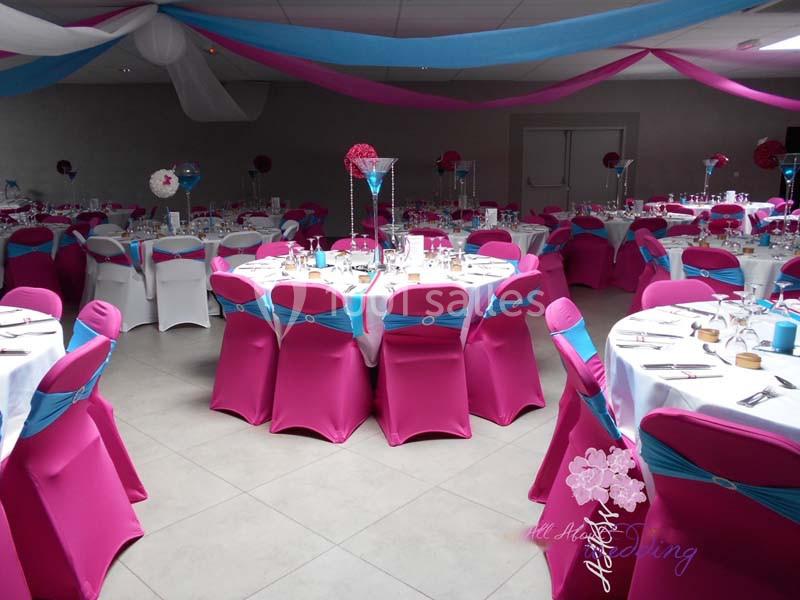 Salle de réception décorée avec des nappes blanches, des chaises roses et bleues, et des centres de table fleuris.